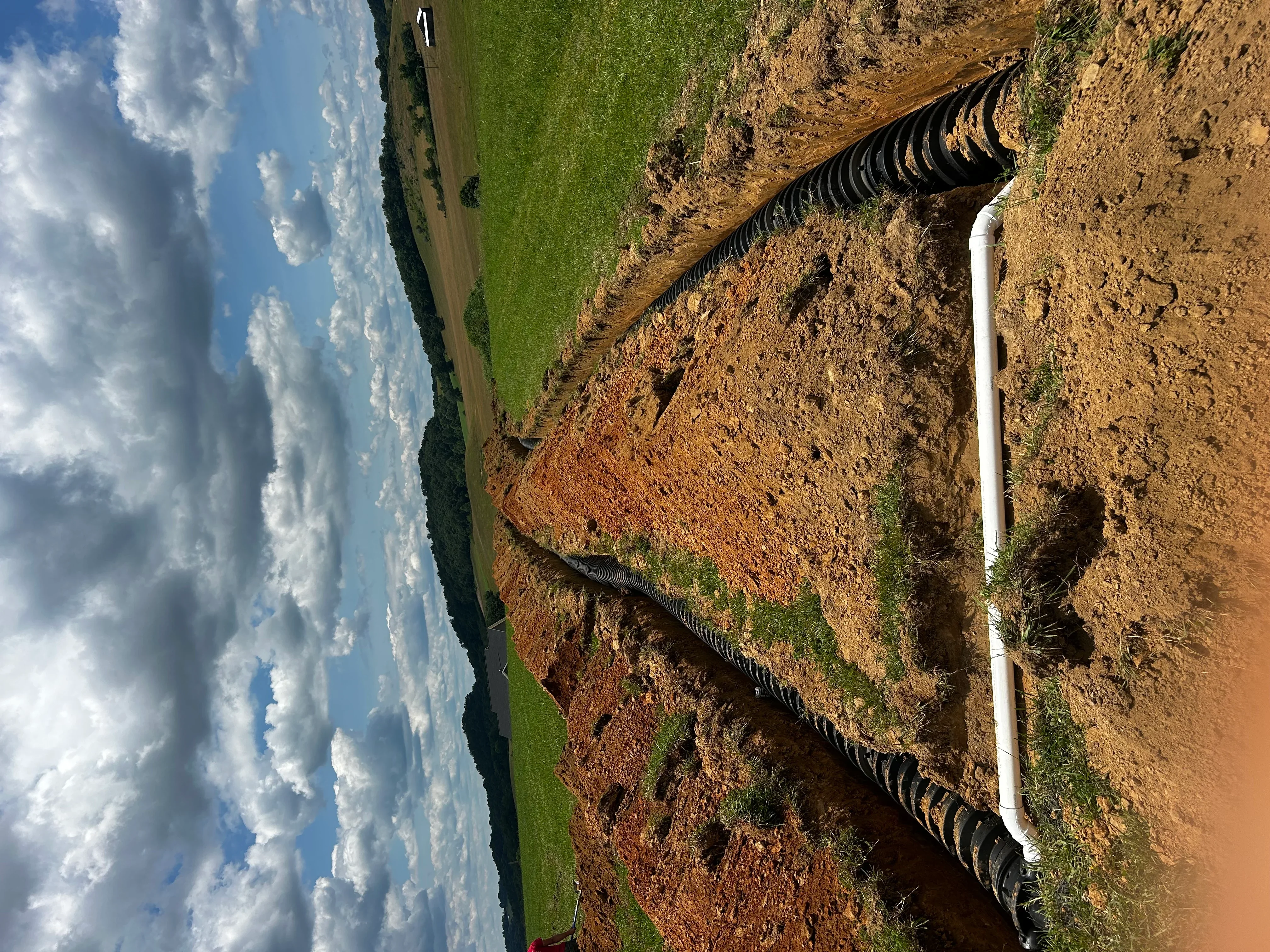 White PVC septic line connected to black corrugated drainage pipes in excavated trenches
