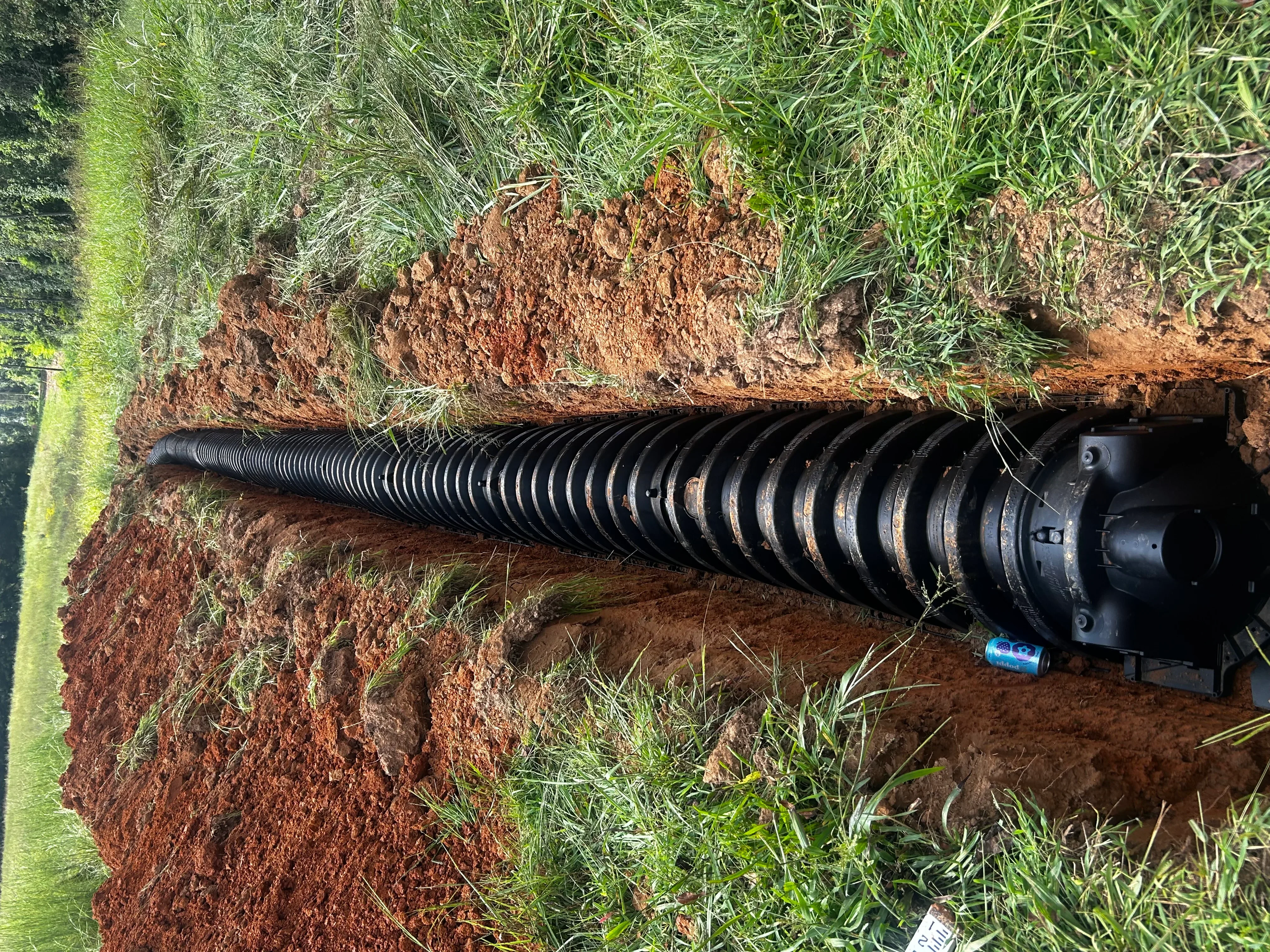 Black corrugated septic tank installed in excavated trench with red clay soil and grass edges