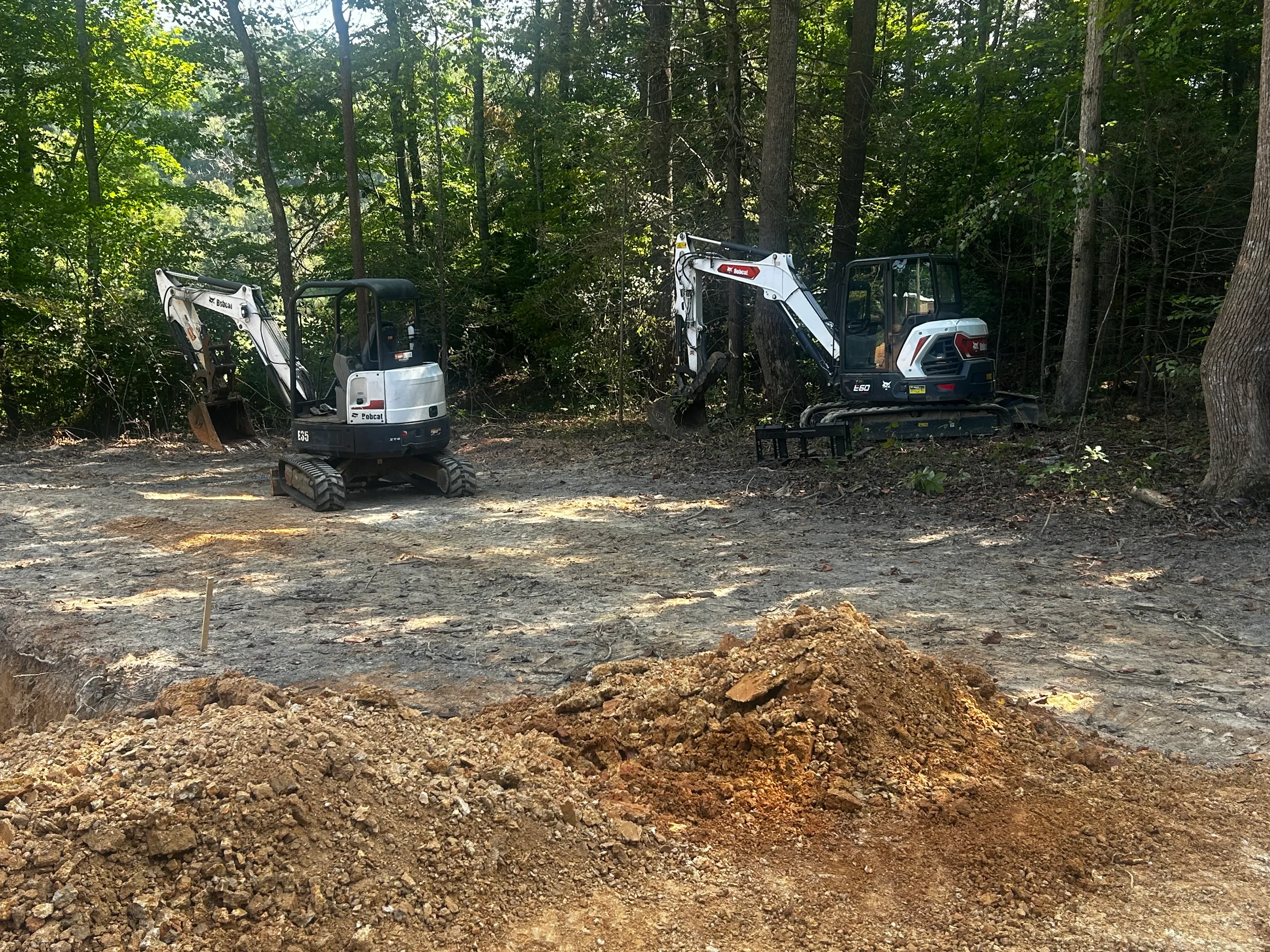 Two compact excavators working on septic system site with excavated soil pile in wooded area