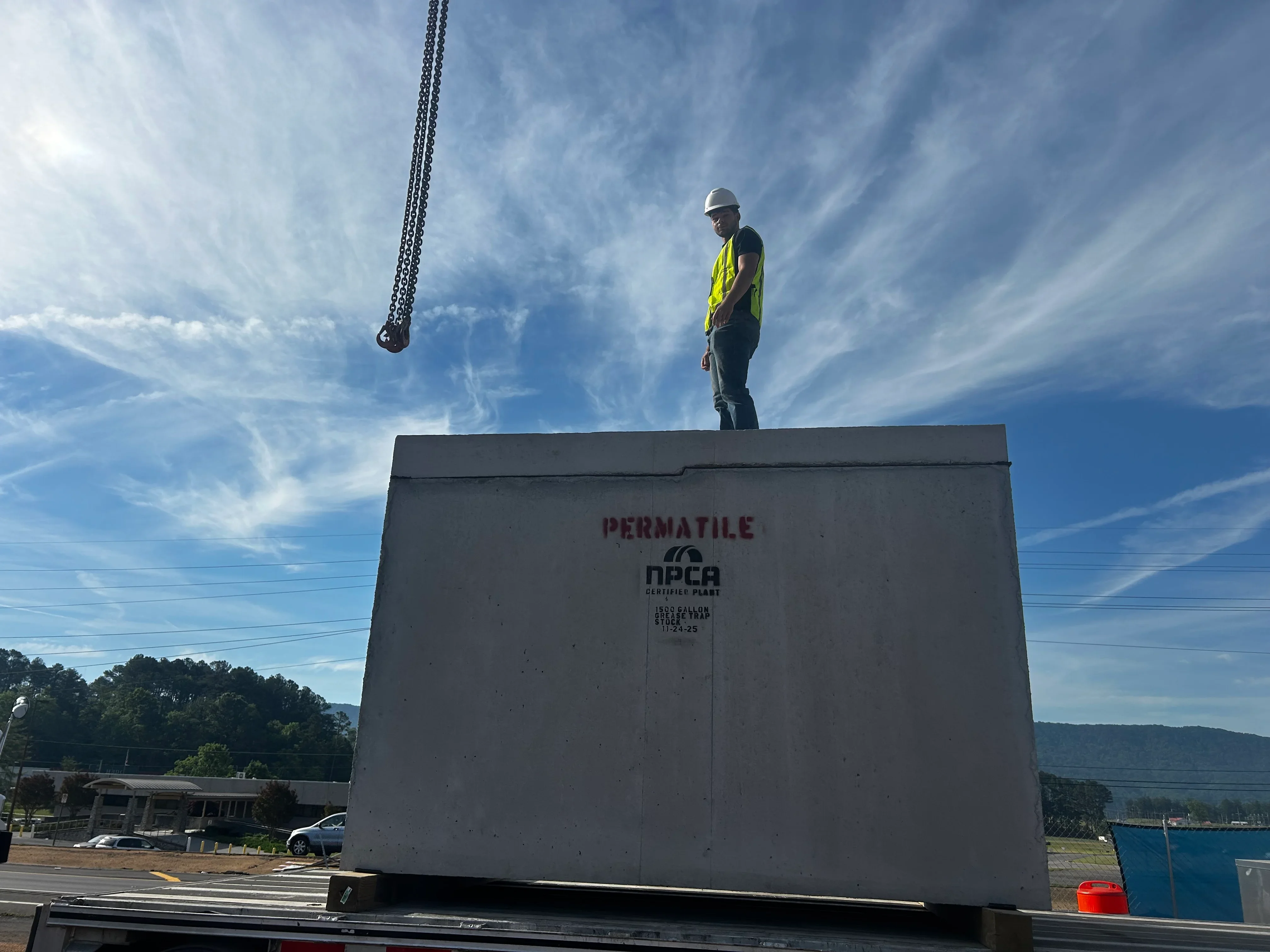 Worker guiding concrete septic tank lowering via chain rigging during installation