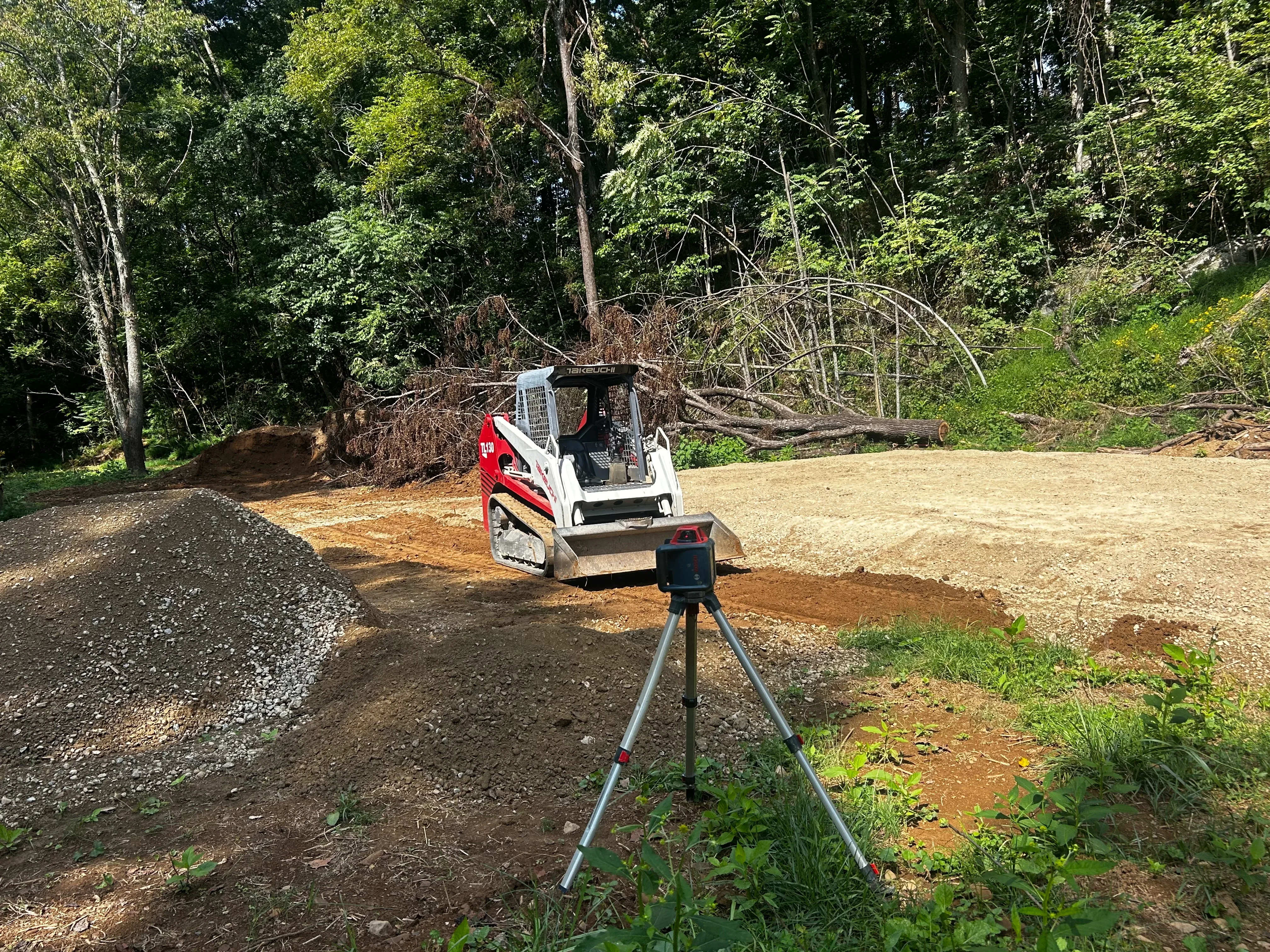Compact track loader grading driveway with stone and soil materials on wooded residential site